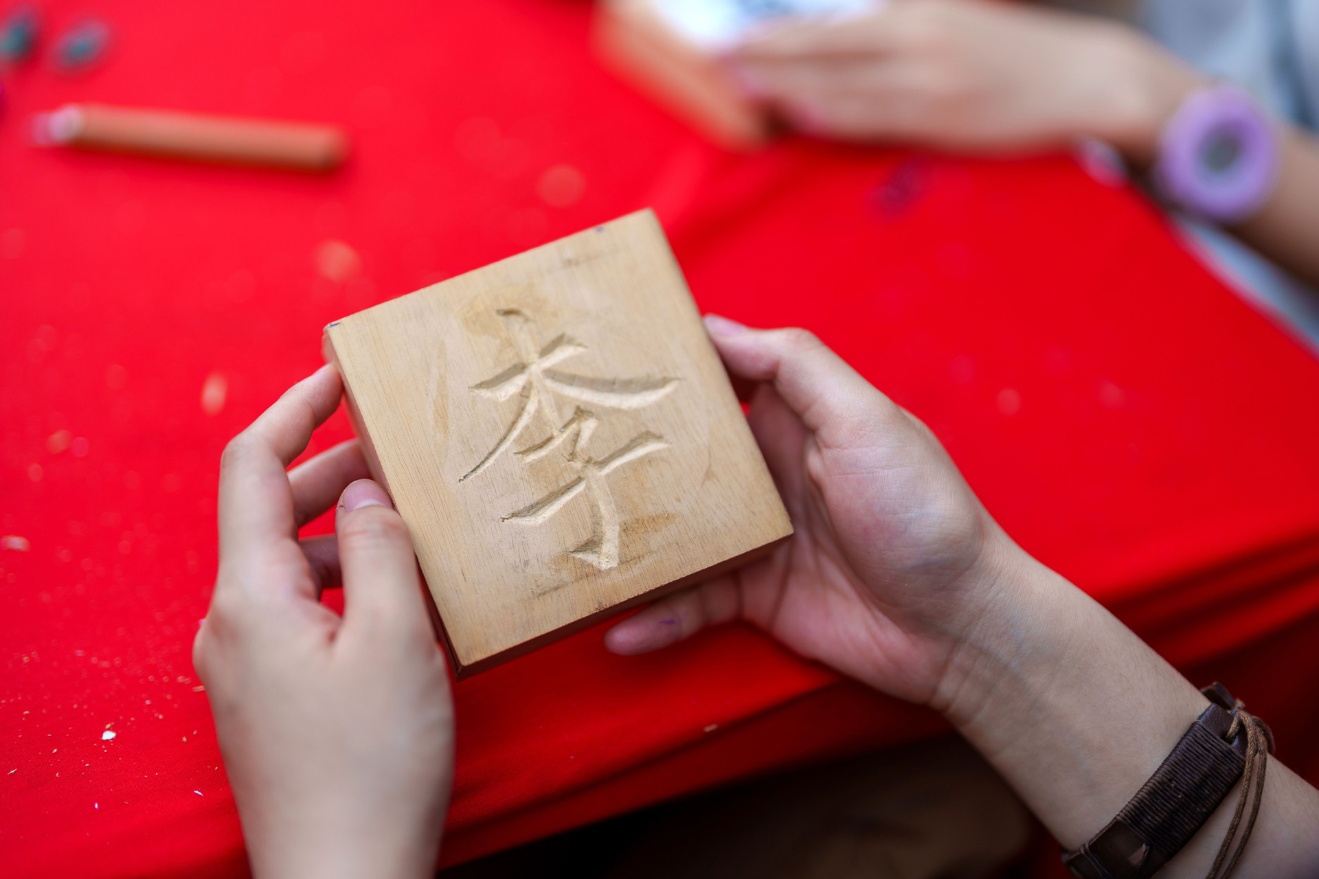 A cropped shot captures a young adult craftsperson meticulously working on a wood carving of a Chinese surname on a wooden block.